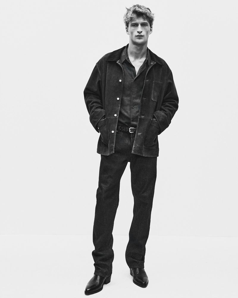 Black and white portrait of a male model wearing a dark jacket, button-down shirt, and trousers against a seamless white background.