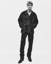 Black and white portrait of a male model wearing a dark jacket, button-down shirt, and trousers against a seamless white background.