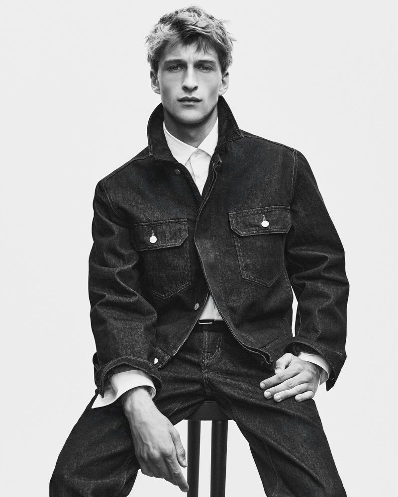 Monochrome portrait of a young male model seated, wearing a dark denim jacket and white collared shirt against a white backdrop.