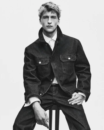 Monochrome portrait of a young male model seated, wearing a dark denim jacket and white collared shirt against a white backdrop.