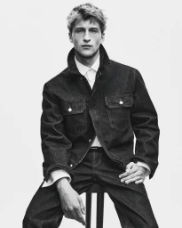 Monochrome portrait of a young male model seated, wearing a dark denim jacket and white collared shirt against a white backdrop.