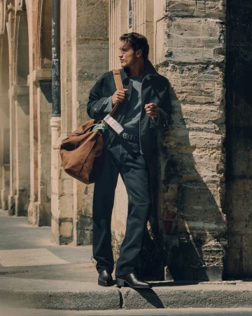 Male model leaning against a stone wall on a sunny street carrying a large brown canvas duffel bag