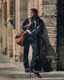 Male model leaning against a stone wall on a sunny street carrying a large brown canvas duffel bag