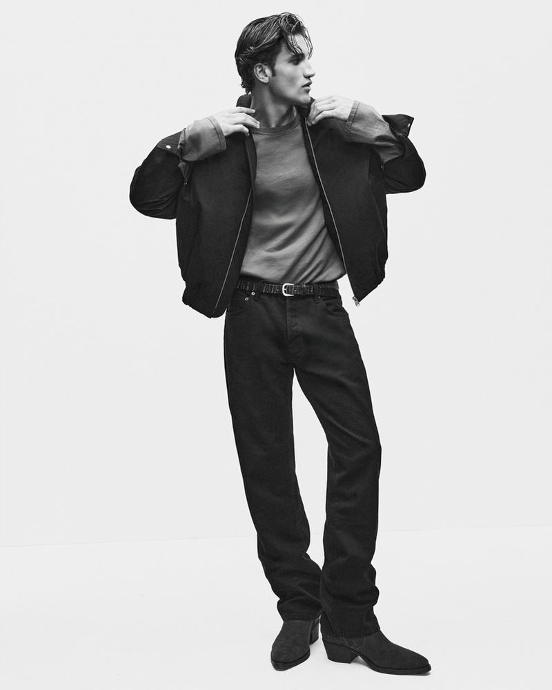 Monochrome full-length fashion portrait of a male model wearing a dark jacket, t-shirt, and jeans against a white backdrop.