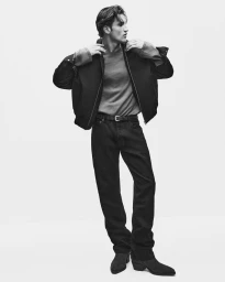 Monochrome full-length fashion portrait of a male model wearing a dark jacket, t-shirt, and jeans against a white backdrop.