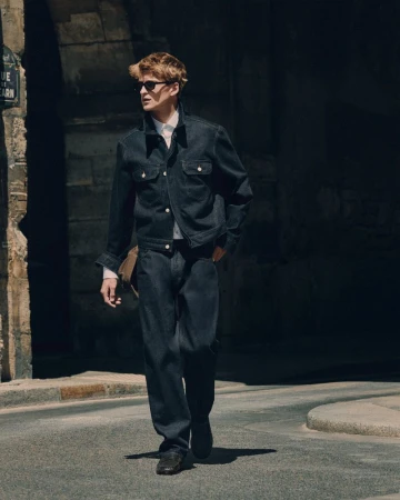 A man wearing a dark denim jacket and matching jeans walks down a sunny urban street.