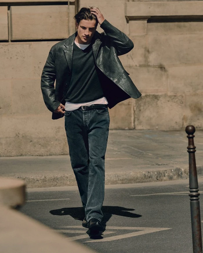 Male model in a dark leather jacket and denim jeans walking past a stone wall exterior.