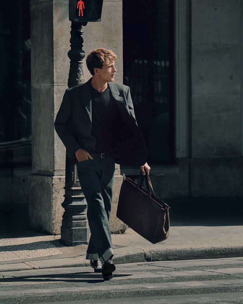 A young male model walking across a city street carrying a large dark leather tote bag wearing a dark blazer and cuffed denim jeans.