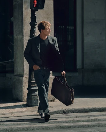 A young male model walking across a city street carrying a large dark leather tote bag wearing a dark blazer and cuffed denim jeans.