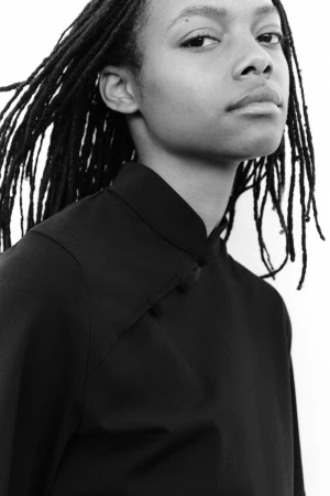 Close-up portrait of a model with long dark dreadlocks wearing a black mandarin collar top against a white background.