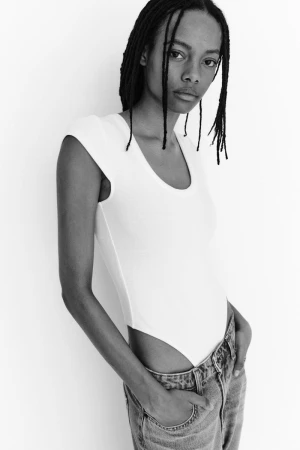 Model with braided hair wearing a white bodysuit and jeans against a white backdrop