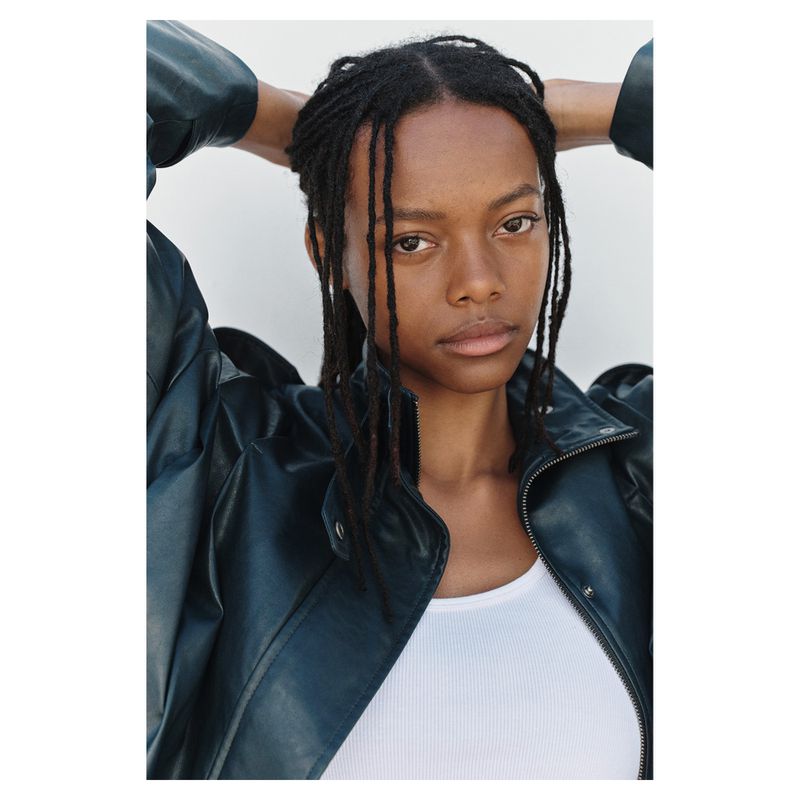 Model with dark braided hair wearing a dark leather jacket and white top against a clean white background