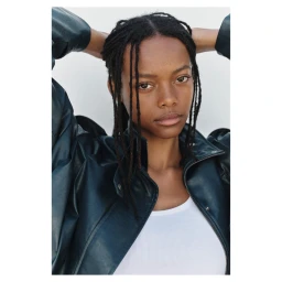 Model with dark braided hair wearing a dark leather jacket and white top against a clean white background
