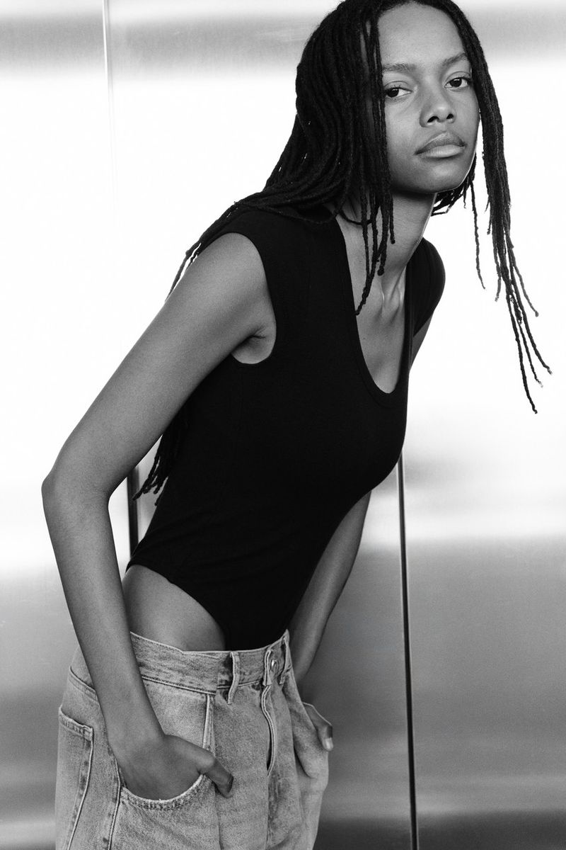 Black and white portrait of a model with braided hair wearing a black sleeveless top and light denim jeans against a metallic background