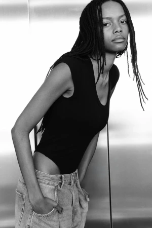 Black and white portrait of a model with braided hair wearing a black sleeveless top and light denim jeans against a metallic background