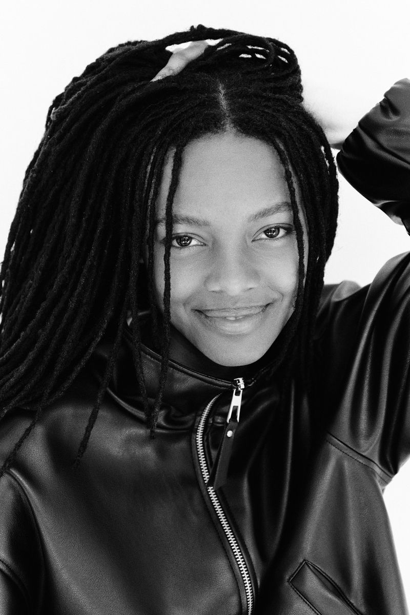 Portrait of a Black woman with long locs wearing a black leather jacket against a plain white background