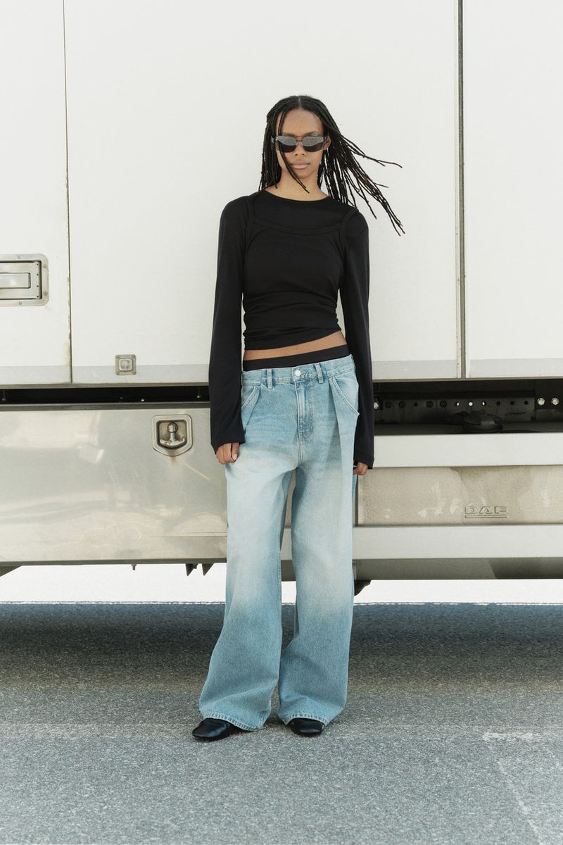 Model wearing dark sunglasses, black long-sleeve top, and light blue denim jeans standing in front of a white truck.