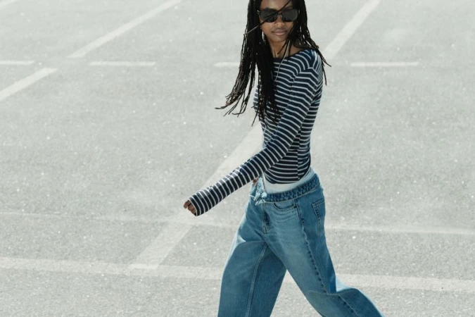 Model wearing dark sunglasses, striped long-sleeve top, and denim jeans walking across an asphalt parking lot surface.