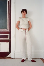 Model standing against a white wall wearing a white top and trousers with a red waist tie and red shoes.