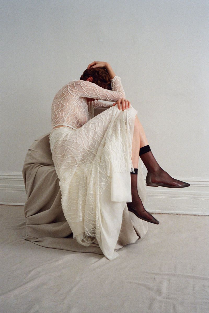 Model seated on draped beige fabric wearing a sheer white patterned long-sleeve top and matching skirt with dark stockings.