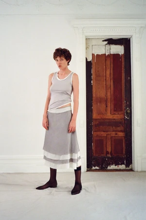 Model wearing a grey tank top and layered skirt standing indoors in front of a weathered wooden door.