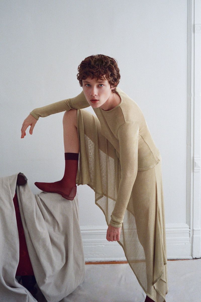 Model Annabelle Weatherly in a long sheer beige outfit with dark red socks posing in a white interior for Mel Usine S/S 27