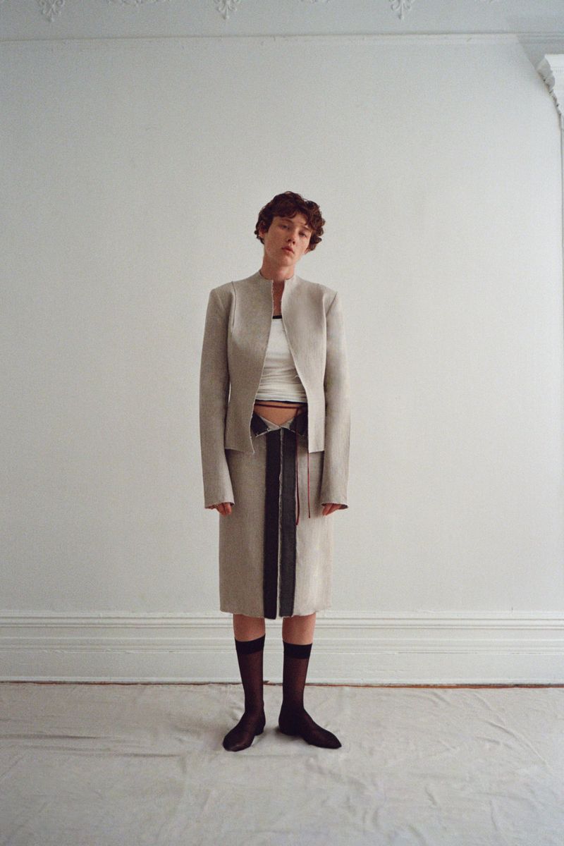 Model wearing a structured beige jacket and skirt set with black sheer knee-high socks against a white interior wall.