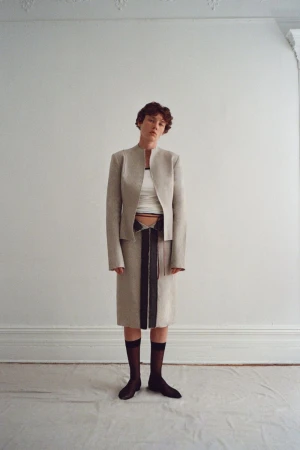 Model wearing a structured beige jacket and skirt set with black sheer knee-high socks against a white interior wall.