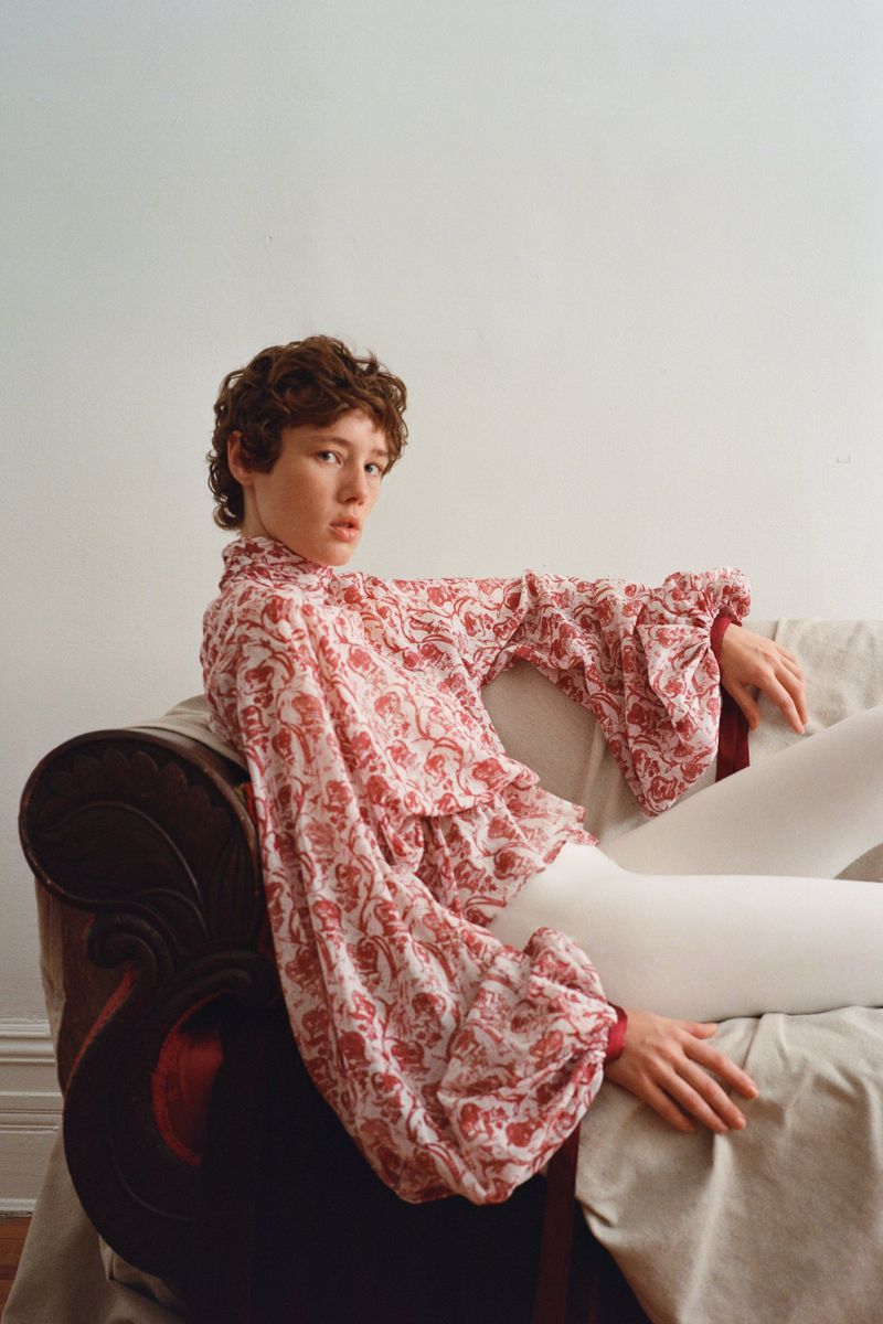 Model Annabelle Weatherly seated on a draped vintage chair wearing a red and white patterned blouse and white trousers
