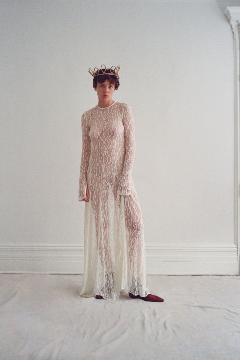 Model wearing a sheer patterned white long-sleeve dress and a metallic crown against a plain white studio background