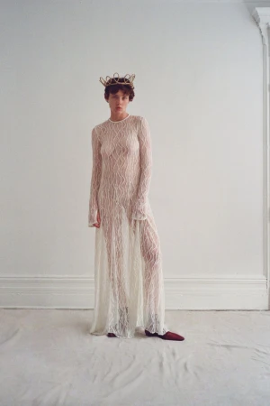 Model wearing a sheer patterned white long-sleeve dress and a metallic crown against a plain white studio background