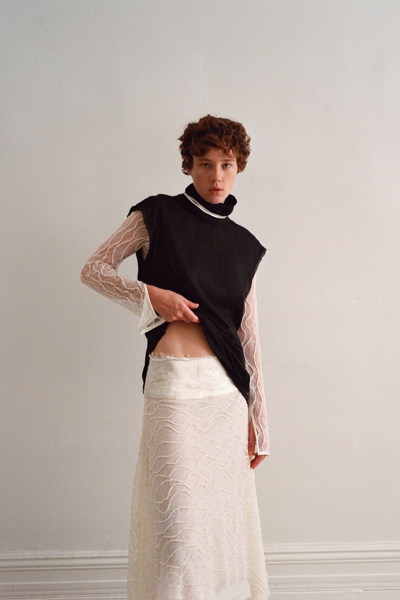 Model with short red hair wearing a black sleeveless top over a sheer white patterned long-sleeve top against a white wall.