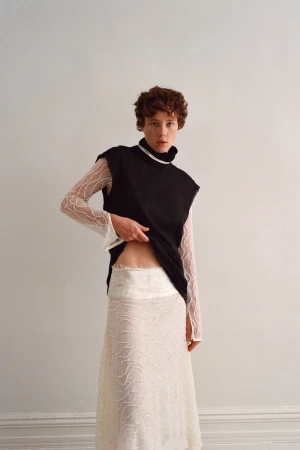 Model with short red hair wearing a black sleeveless top over a sheer white patterned long-sleeve top against a white wall.