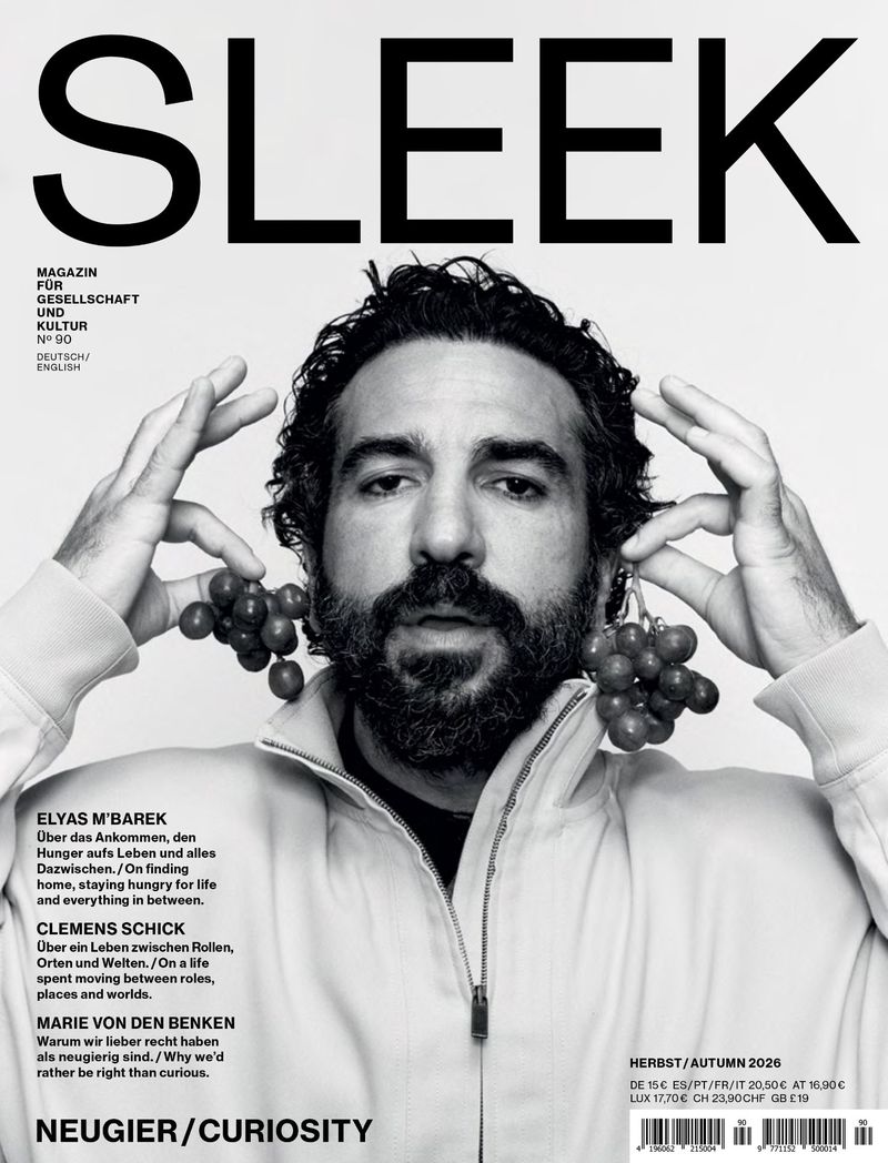 Close-up magazine cover portrait of a bearded man holding two clusters of red grapes next to his ears against a light background.