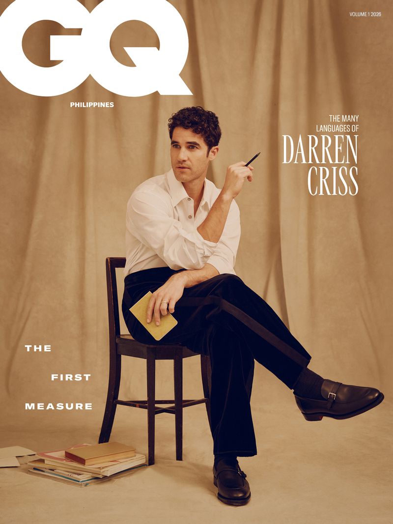 Darren Criss seated on a wooden chair holding a pen and notebook against a draped neutral fabric backdrop