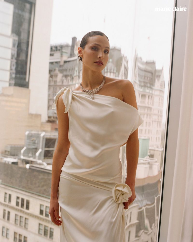 Model wearing a white one-shoulder dress and diamond necklace standing before a large window with city buildings outside.