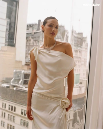 Model wearing a white one-shoulder dress and diamond necklace standing before a large window with city buildings outside.