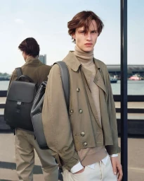 Young man wearing a tan jacket and backpack outdoors by the waterfront for RIMOWA
