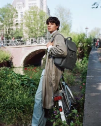 Young man in a beige trench coat standing beside a bicycle on an Amsterdam canal bridge carrying a grey backpack