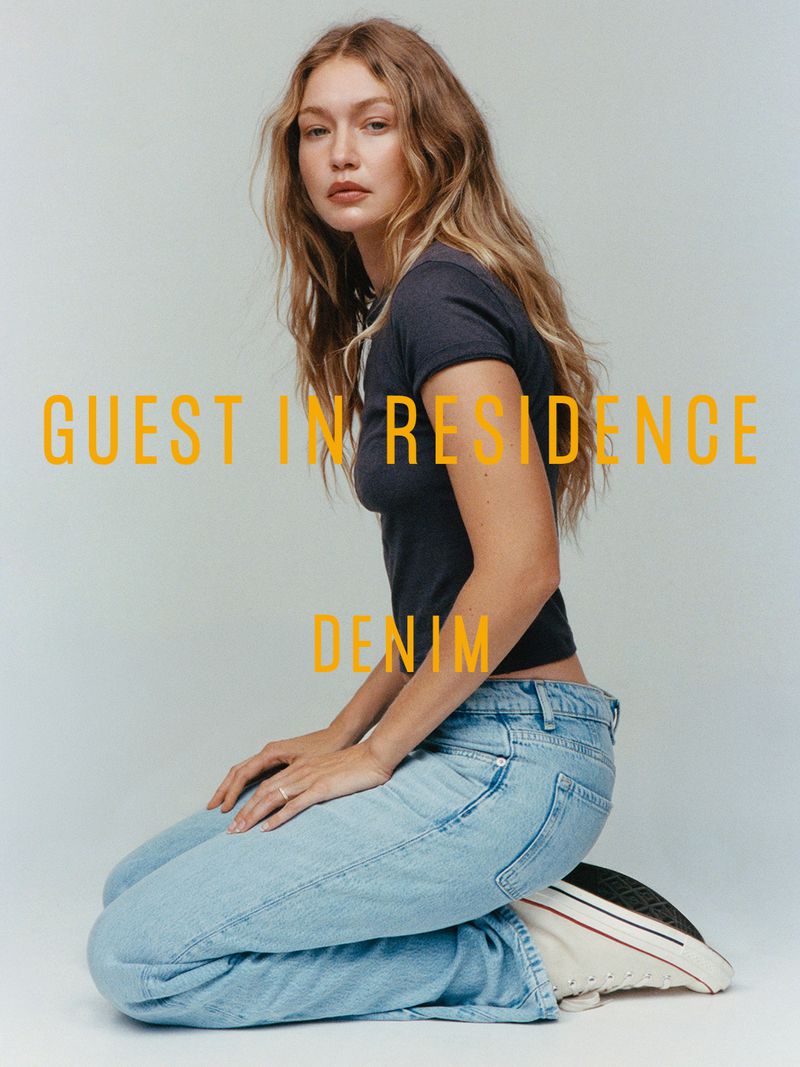 Gigi Hadid kneeling wearing a dark t-shirt and light blue jeans against a grey studio backdrop