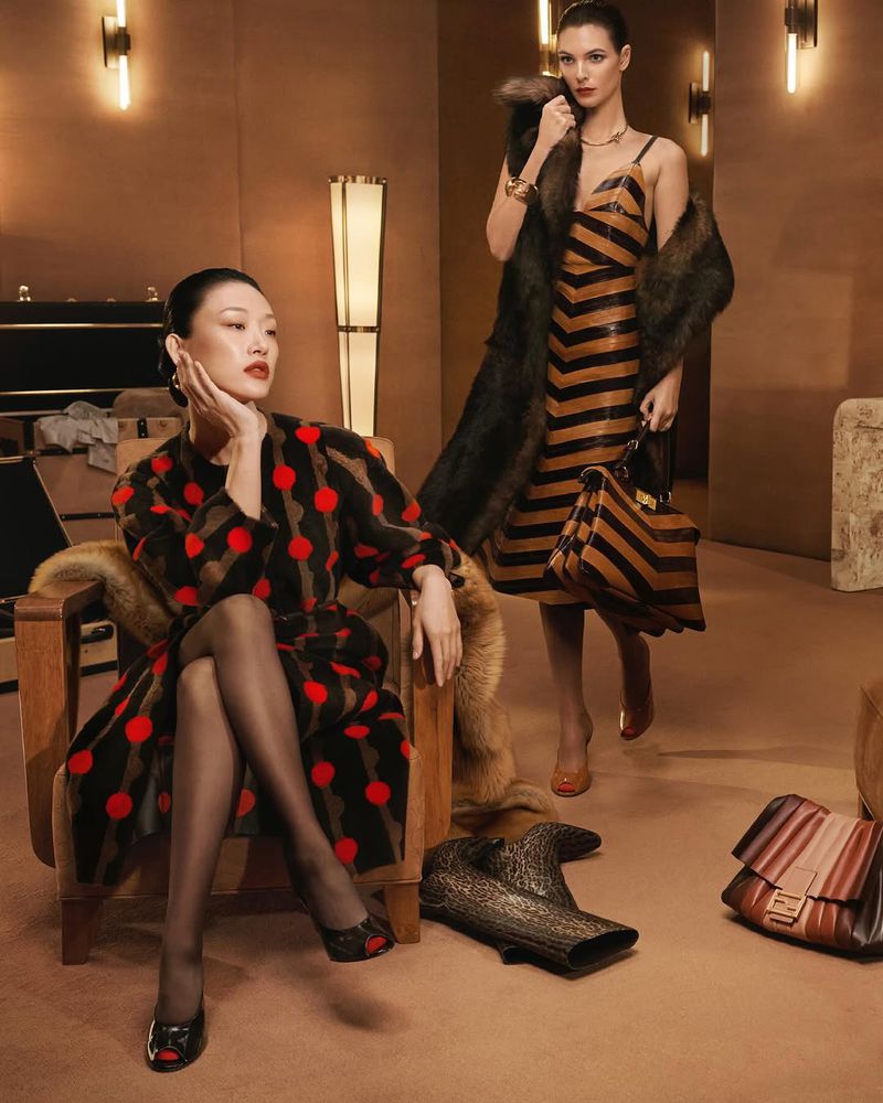 Two women in stylish vintage-inspired dresses pose in a warm-toned studio setting, complemented by retro furniture and designer handbags and boots.