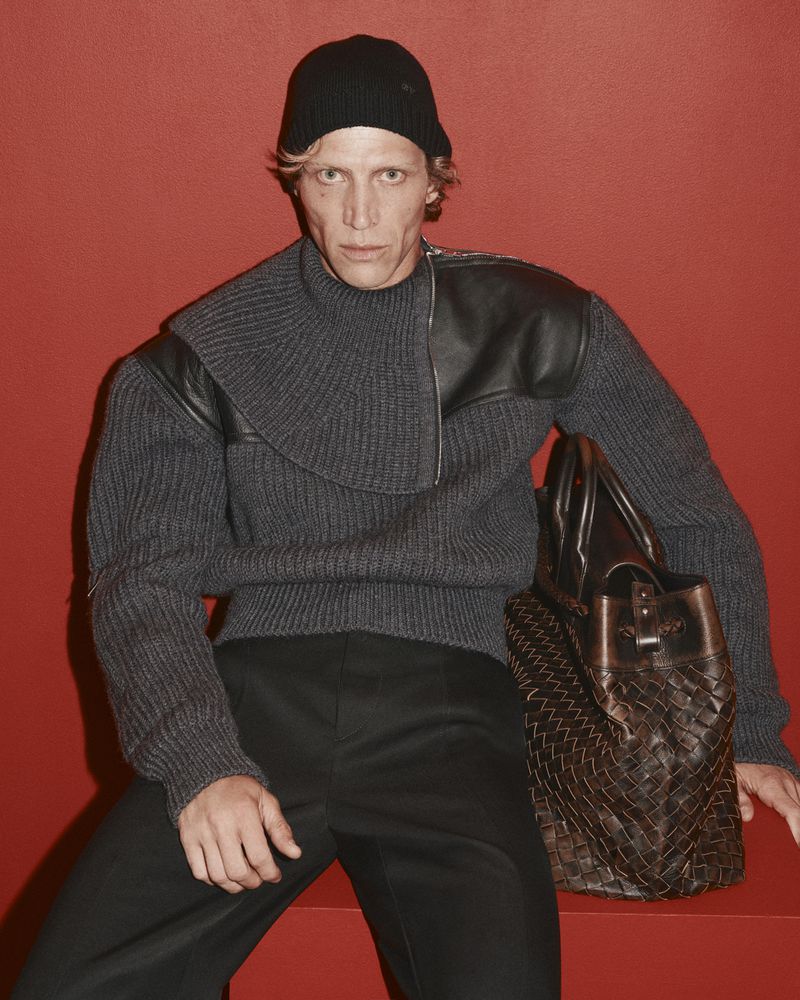 Male model wearing a grey sweater and black beanie holding a woven leather bag against a red background