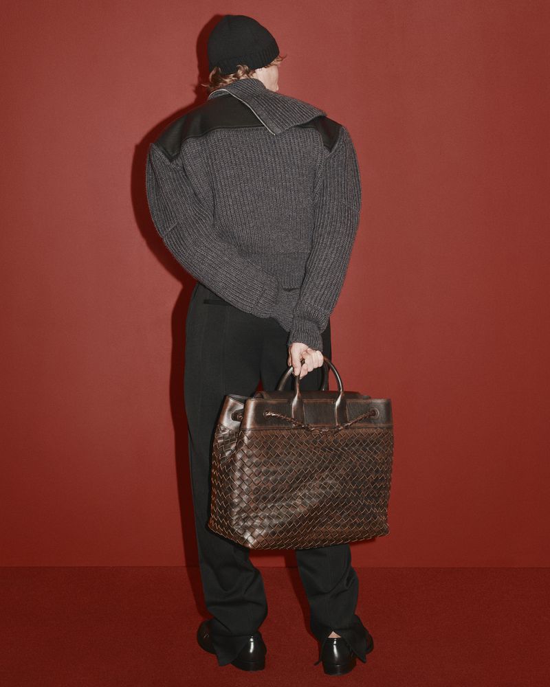 Model wearing dark ribbed sweater, black beanie and trousers, holding a large brown leather woven bag against a solid red background.
