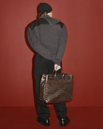 Model wearing dark ribbed sweater, black beanie and trousers, holding a large brown leather woven bag against a solid red background.