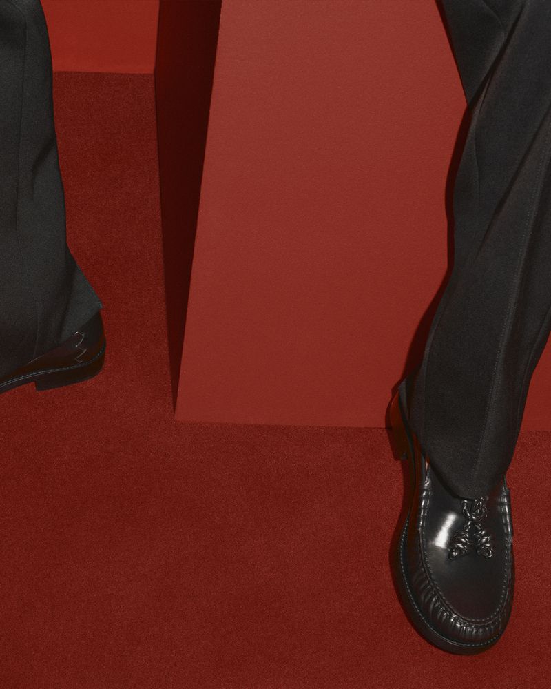 Close-up of a person wearing black trousers and leather loafers standing on a vibrant red carpet against a matching red studio backdrop.