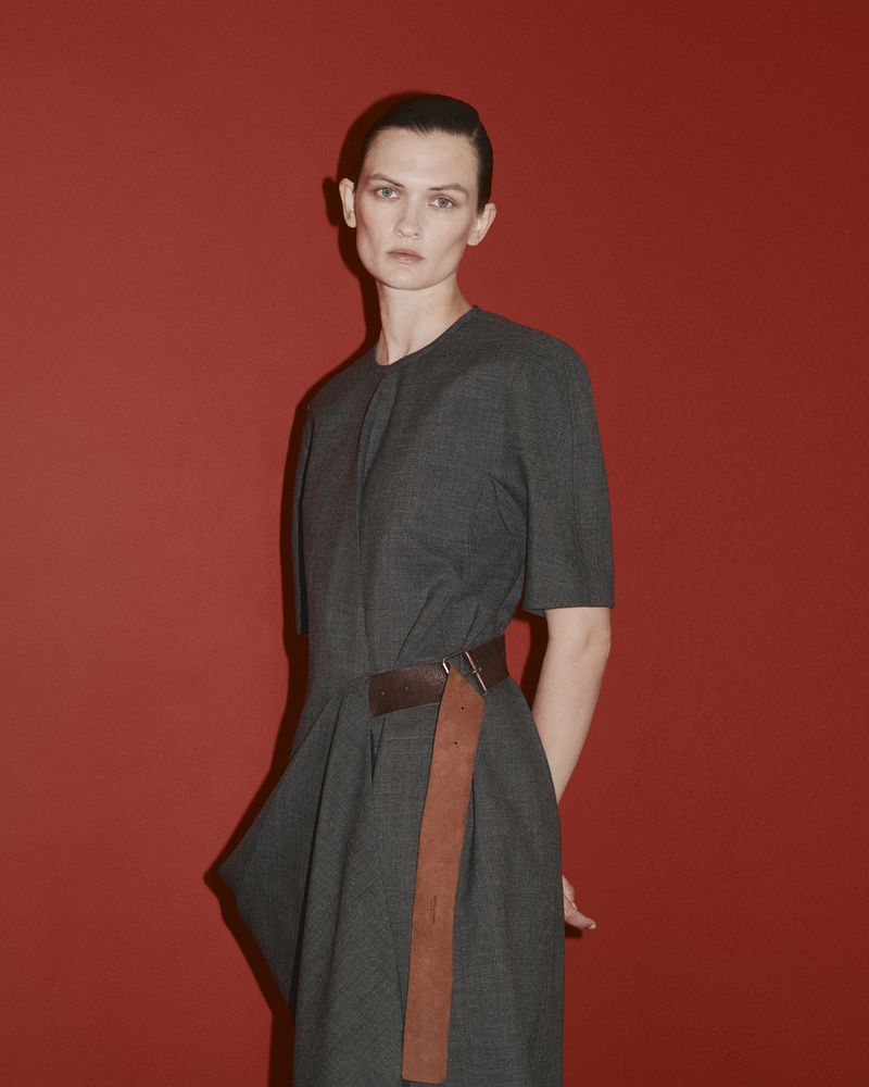 Model Lara Mullen wearing a grey dress with a brown belt against a red background for Bottega Veneta