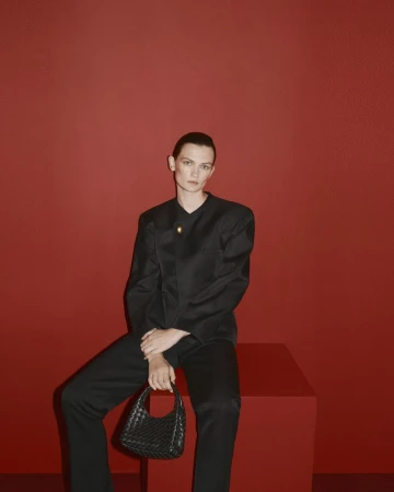 Model Lara Mullen seated on a red block wearing a black jacket and holding a black woven handbag against a solid red studio background.