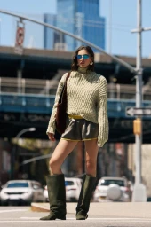Model wearing a textured green knit sweater, shorts, and tall boots standing on an urban street near a bridge.