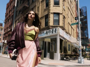 Model standing on a city street wearing a green camisole, pink skirt, and maroon bomber jacket holding a small patterned handbag.