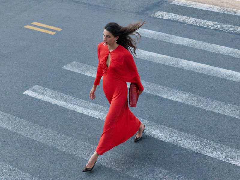 Model wearing a vibrant red dress walking across an asphalt crosswalk holding a red clutch
